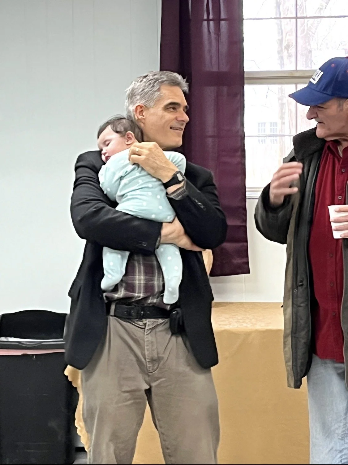 A man holding a sleeping baby, standing indoors with another man wearing a baseball cap and jacket, near a window and a purple curtain.