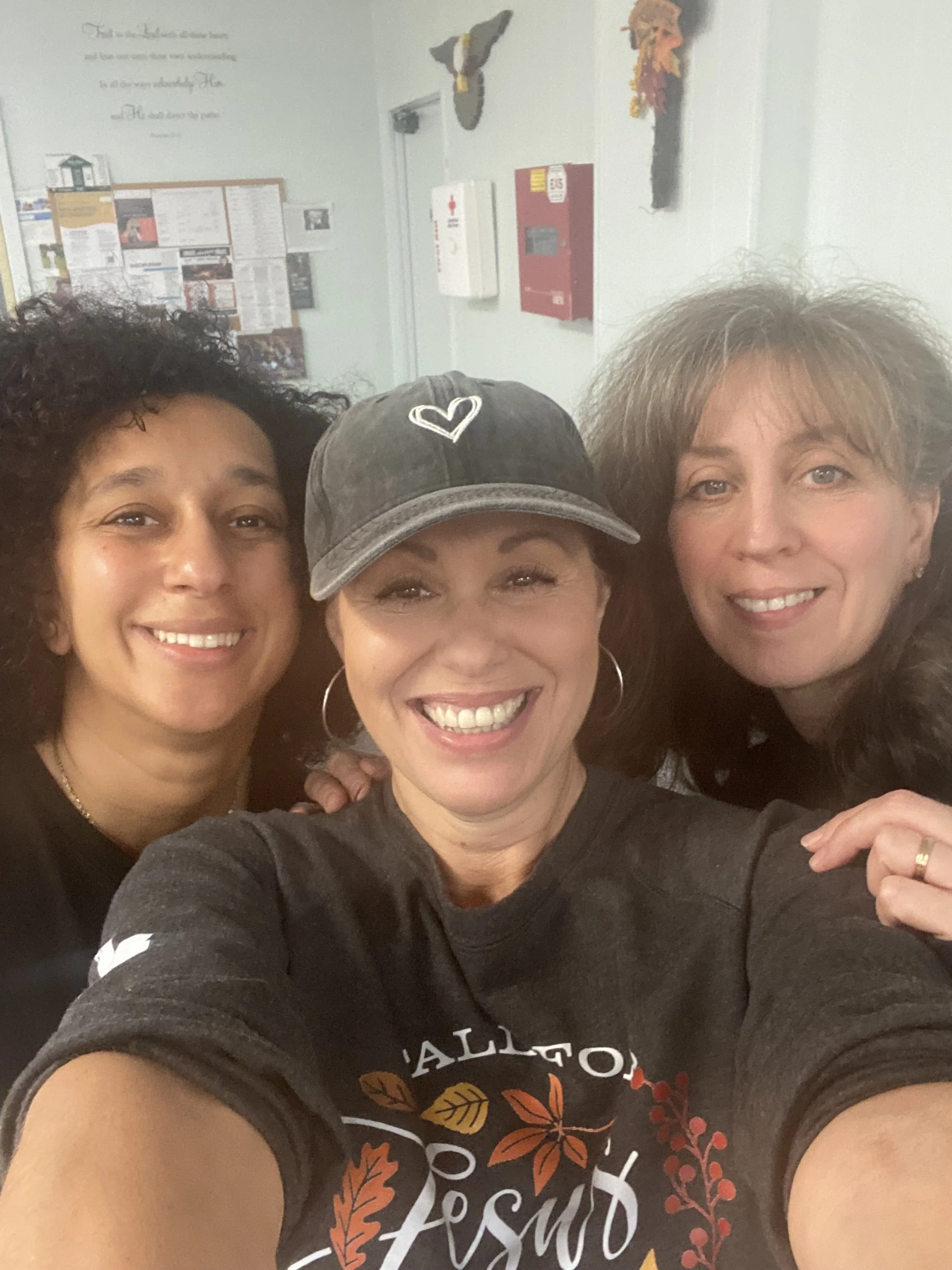 Three women smiling closely together indoors, with bulletin boards and safety equipment visible on the wall behind them.