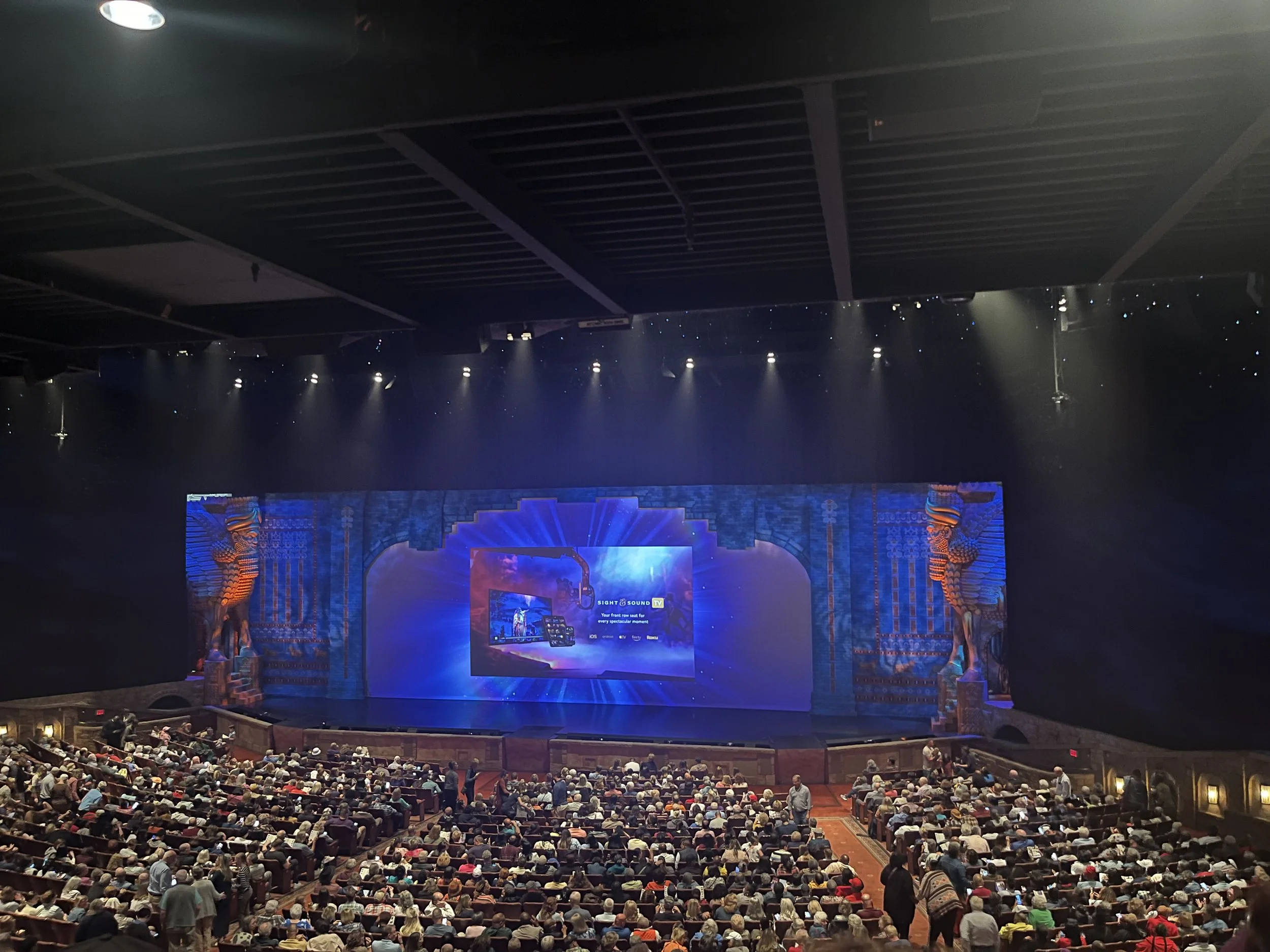 Wide shot of a theater filled with an audience, facing a large stage with a vibrant blue and purple backdrop, featuring intricate designs and statues of lions on each side.