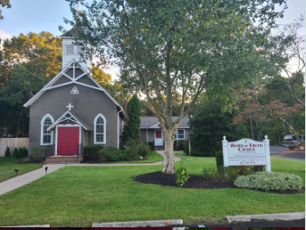 A church with a gray exterior, white trim, and a red door, surrounded by a well-maintained lawn and trees, with a sign in front.