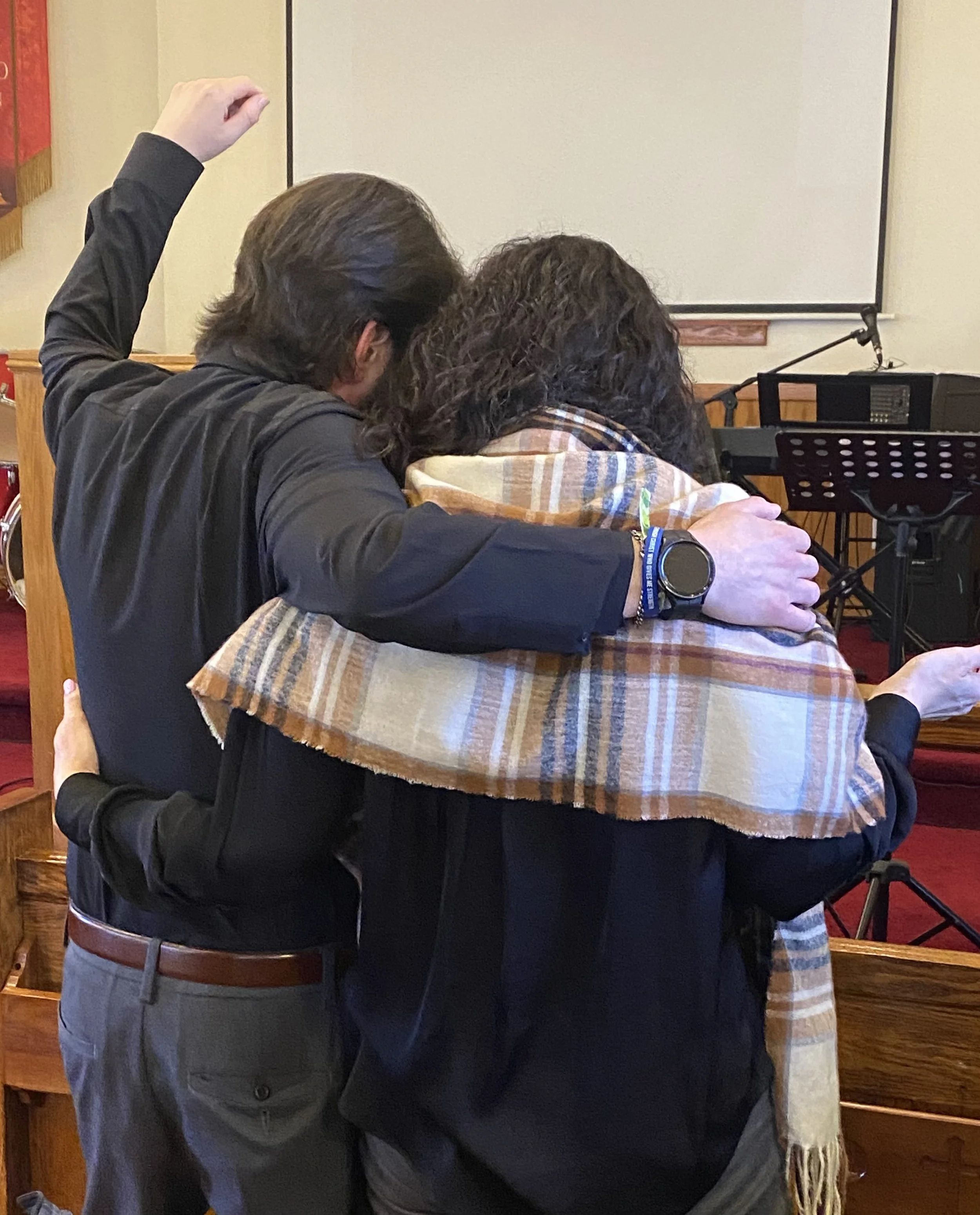 Two people embracing in a church or community setting, one with a plaid shawl draped over the other's shoulders, and a band on the person's wrist.