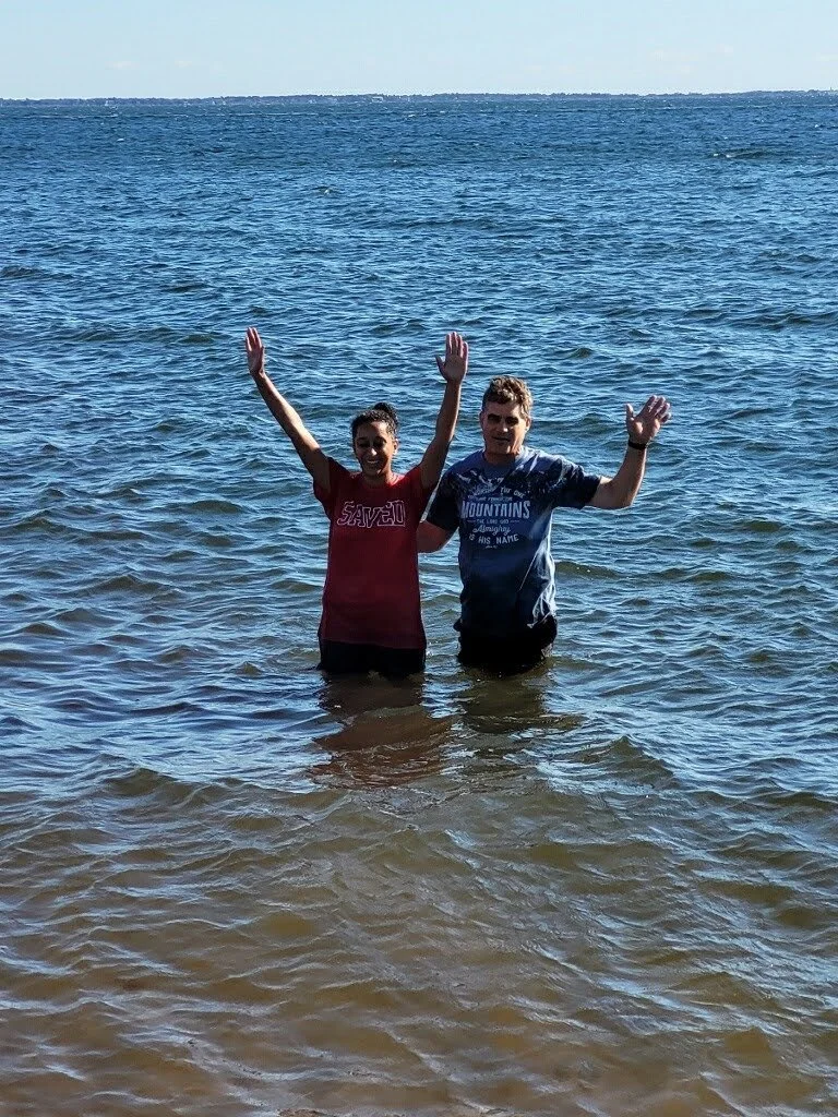Two people standing in the ocean water with their hands raised, smiling, under a clear blue sky.