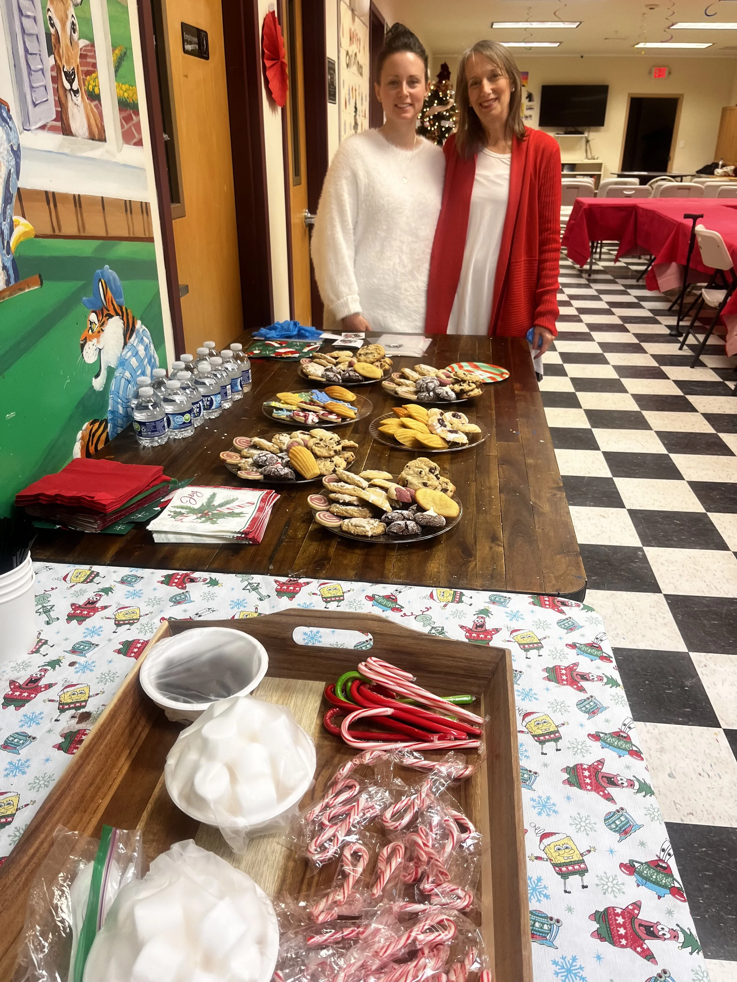 Two women stand behind a table with various cookies and treats at a holiday event. The background includes a decorated Christmas tree and a decorated room with red tablecloths and checkered floor.