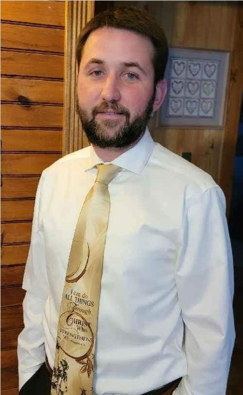 A man with dark hair and a beard wearing a white dress shirt and a cream-colored tie with religious text, standing in front of wooden walls and framed heart artwork.
