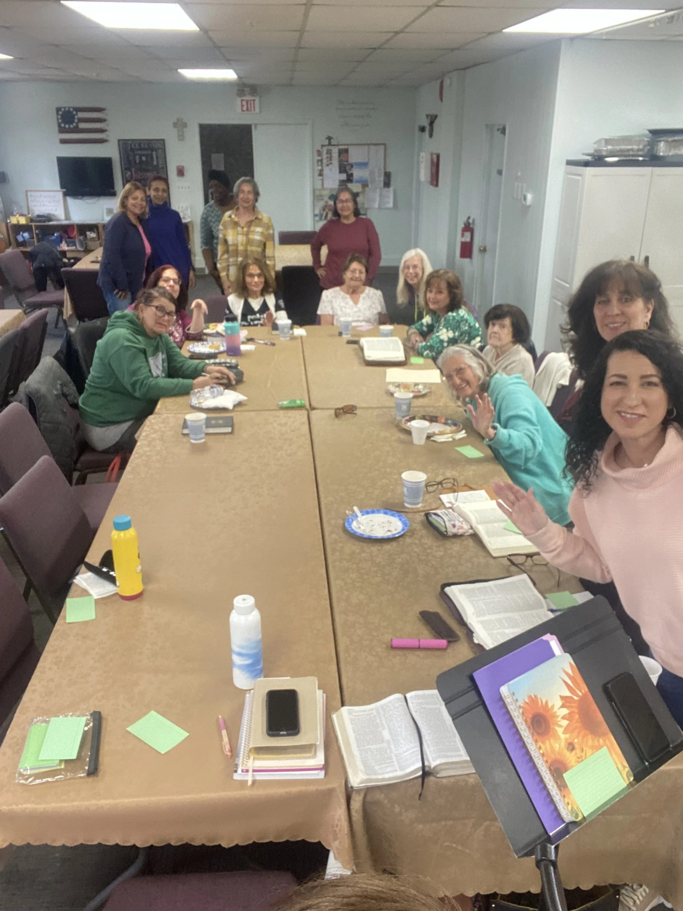 Group of women gathered around a large conference table in a room decorated with American flag, a cross, and various notices pinned on the wall, some women are sitting and some are standing, with books, notebooks, cups, and other items on the table.