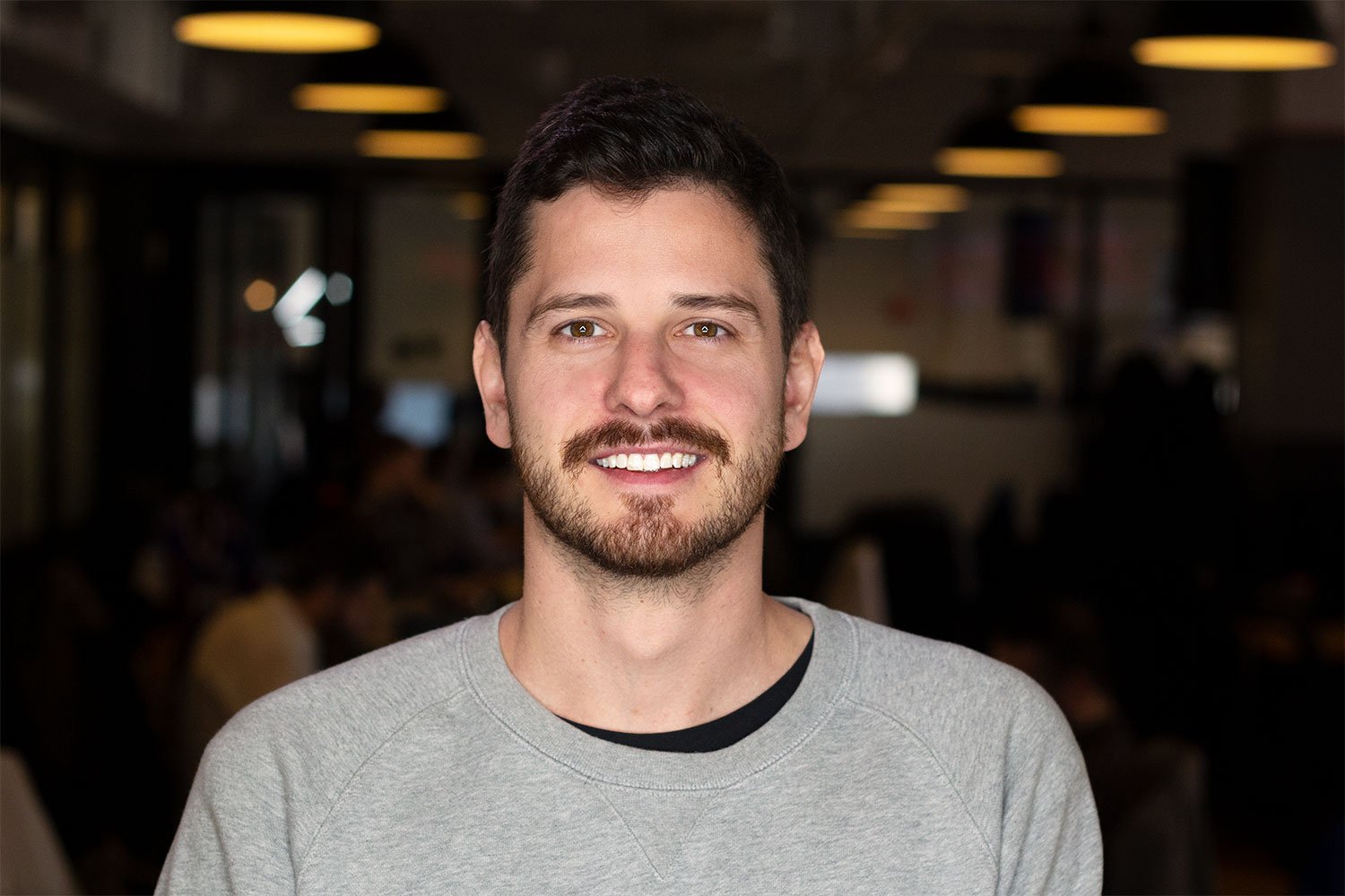 A young man with dark hair, beard, and mustache smiling at the camera in an indoor setting with dim lighting and blurred background.