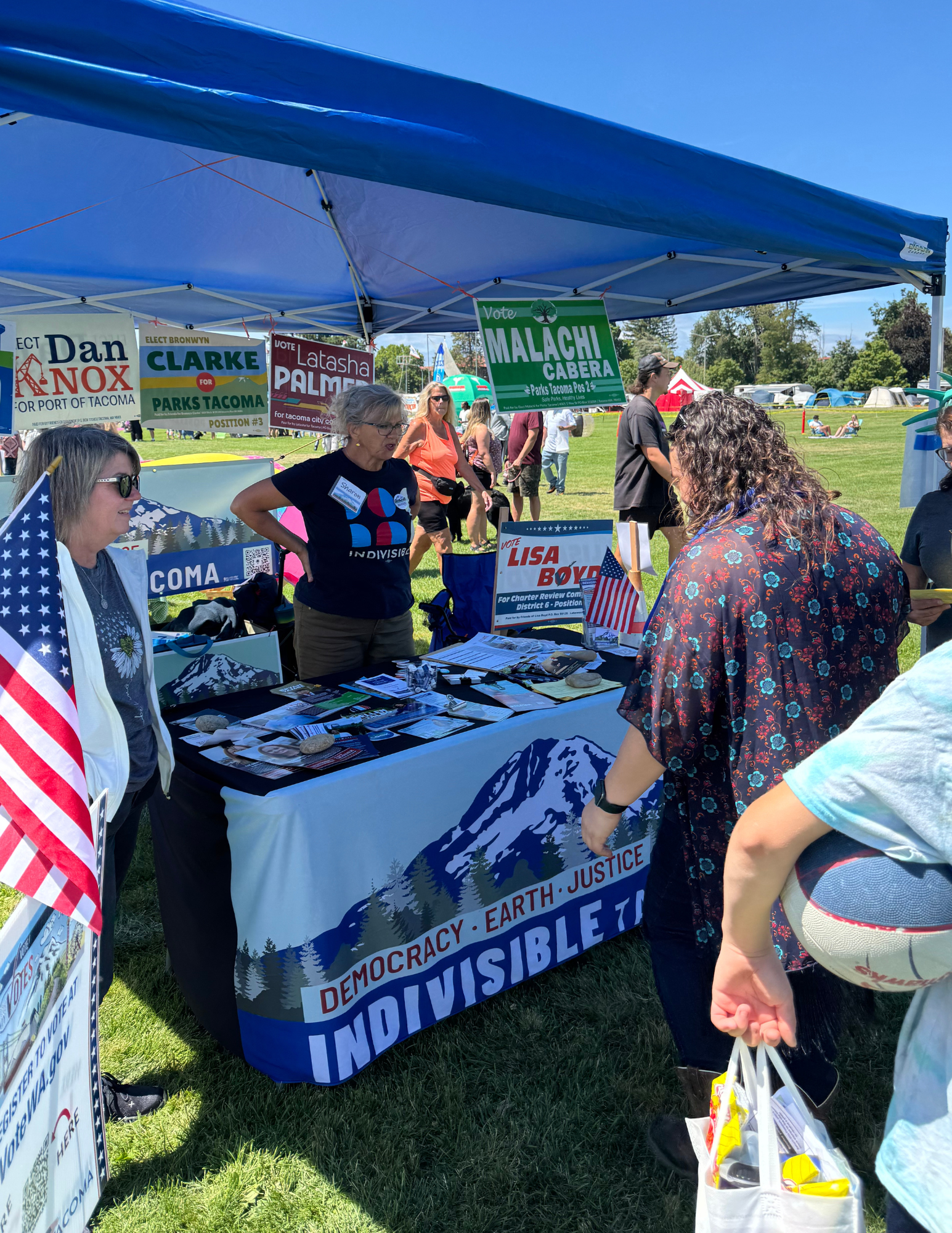 People visiting the Indivisible Tacoma booth at an outdoor event with banners for candidates and issues, and a table displaying informational materials.