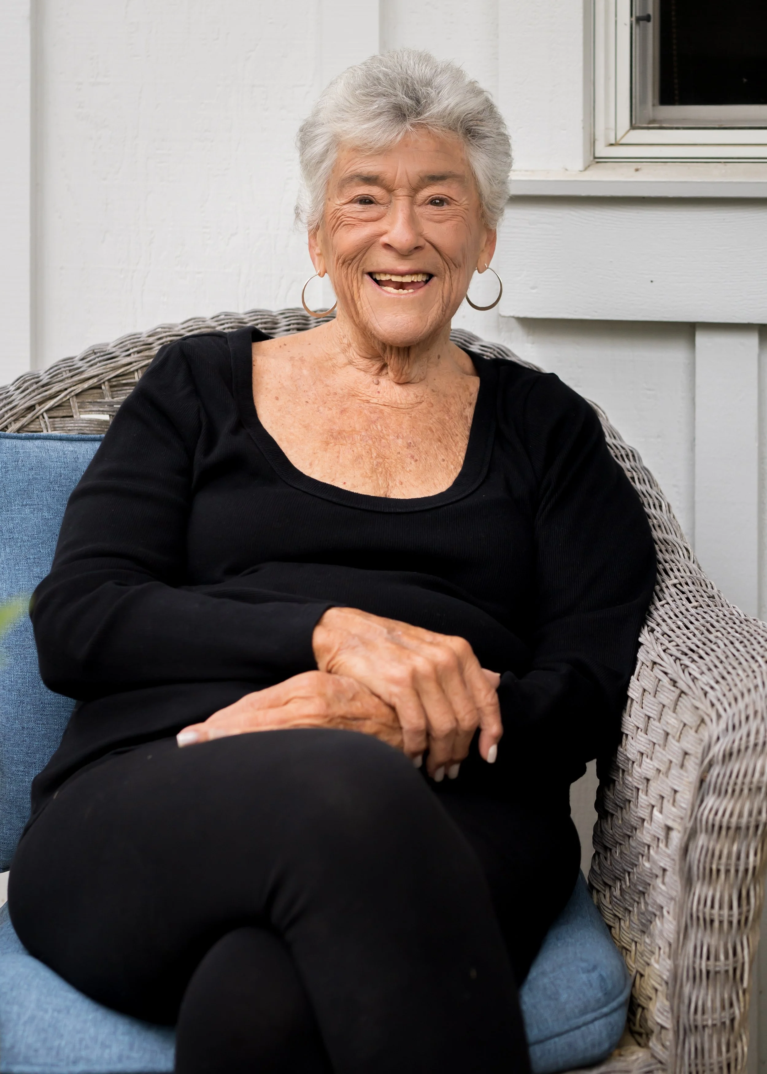 An elderly woman with short gray hair, wearing hoop earrings and a black long-sleeve top, sitting on a wicker chair with a blue cushion, smiling outdoors near a building.