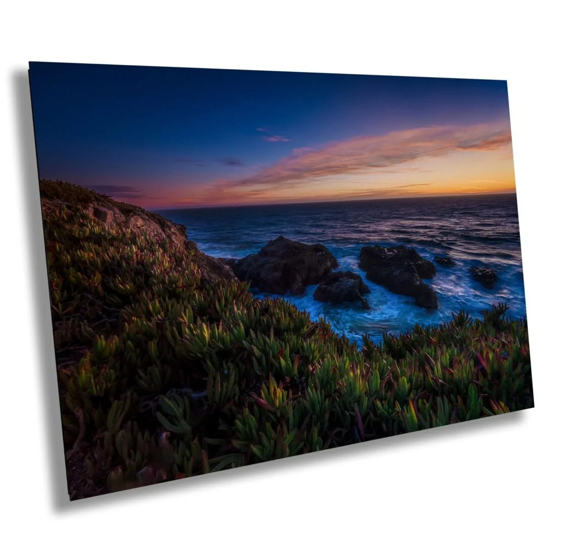 A coastal landscape at sunset with a rocky shoreline, ocean waves, and a colorful sky with pink and orange clouds.