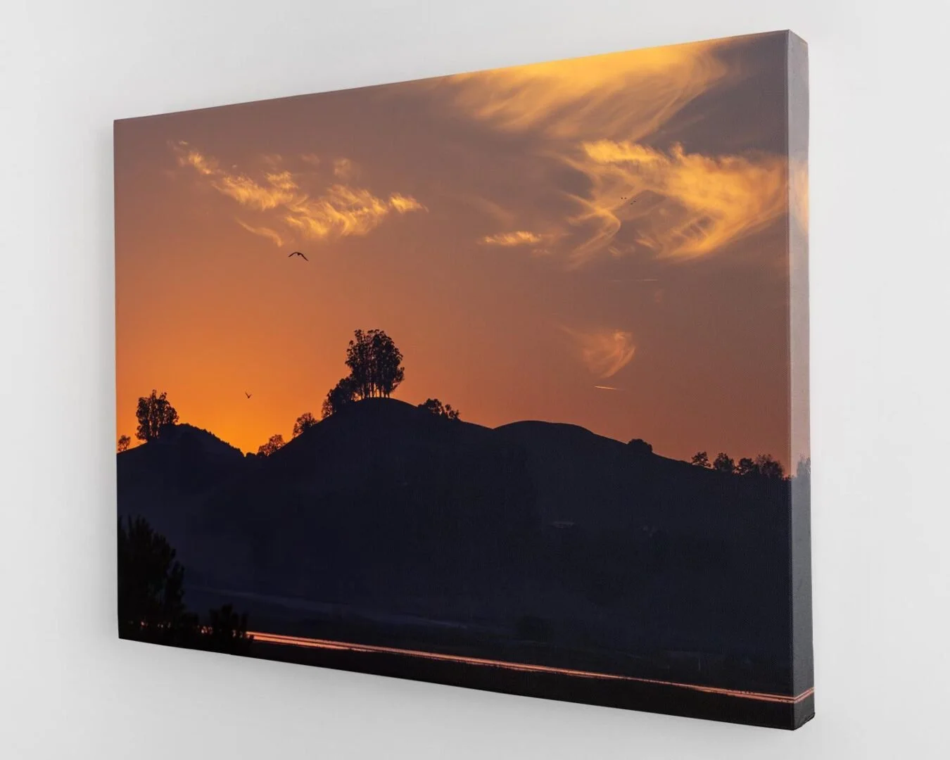 Sunset over hills with silhouetted trees and birds flying in the sky, warm orange and yellow hues in the clouds.