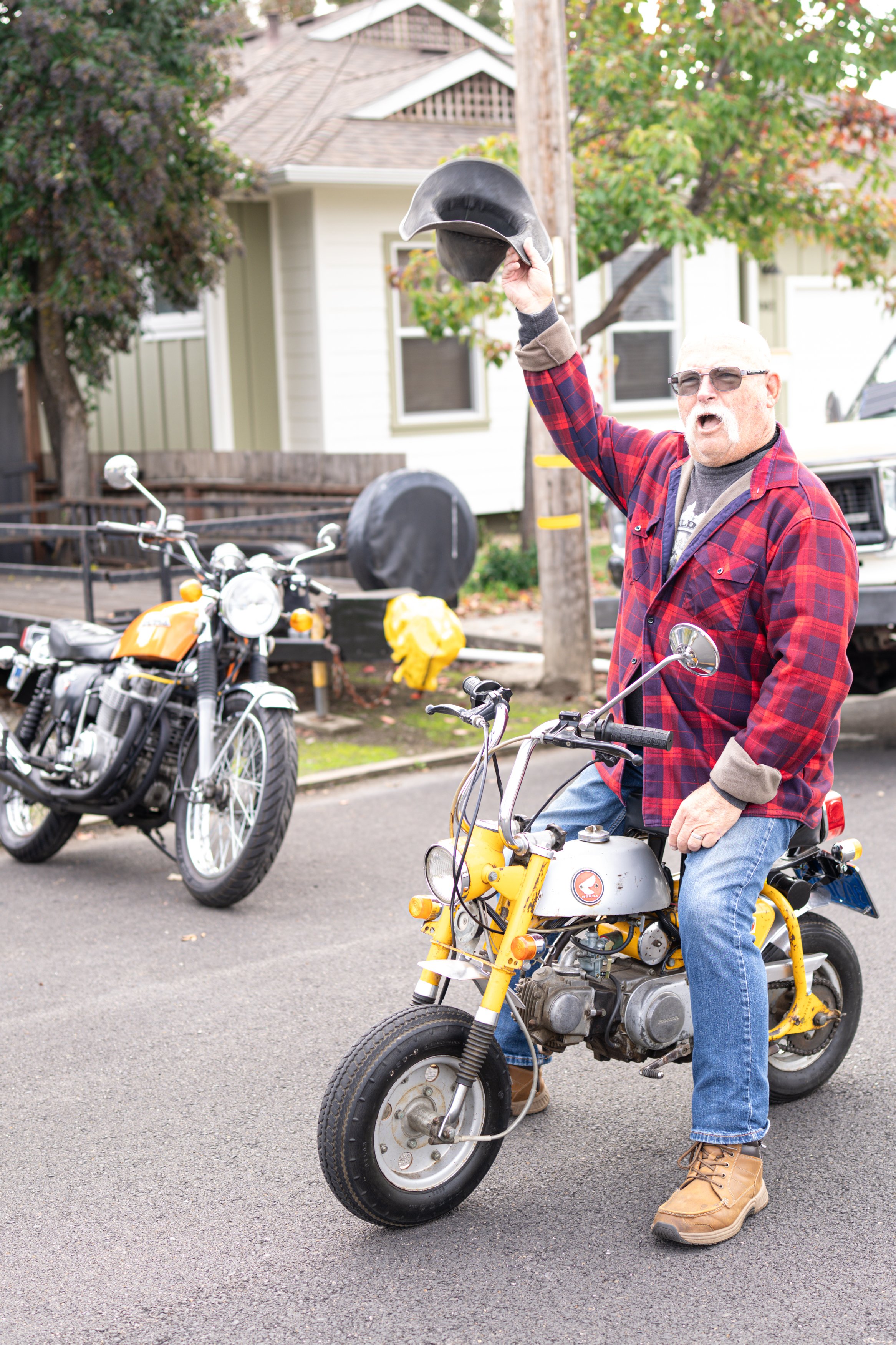 A man wearing sunglasses, a red plaid shirt, and brown boots sitting on a small yellow motorcycle, holding a black helmet in the air, with a black and orange motorcycle parked behind him.