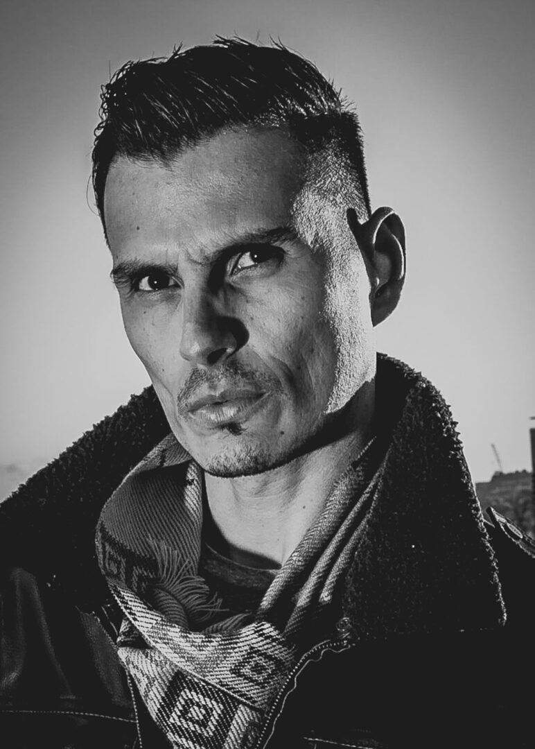 Black and white close-up portrait of a man with styled hair, wearing a scarf and jacket, with a serious expression.