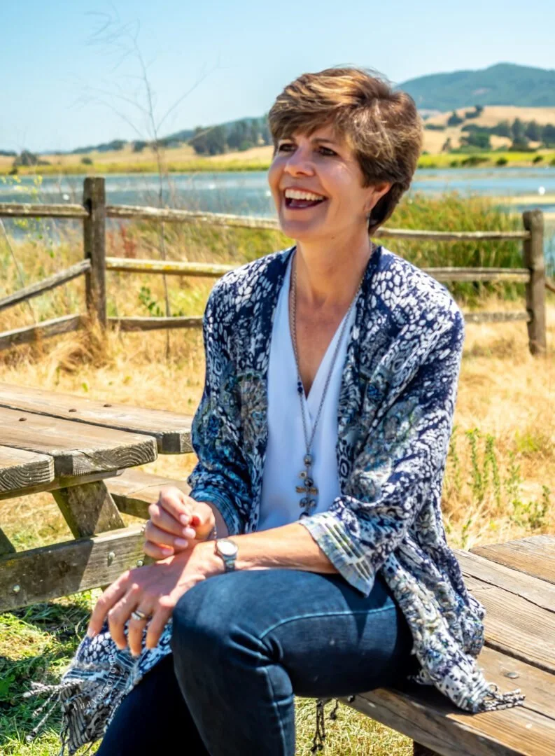 A woman with short brown hair laughing outdoors, sitting on a picnic table near a fence with a lake and rolling hills in the background.