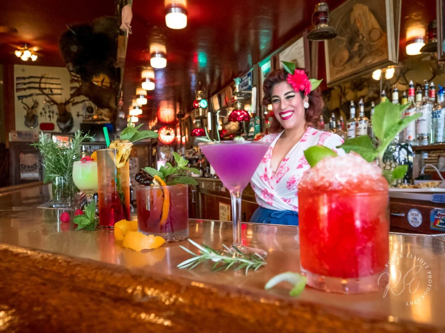 A cheerful woman with red hair and a flower in her hair, smiling behind a bar with colorful cocktails and garnishes, in a cozy, rustic bar with deer antlers on the wall and warm lighting.
