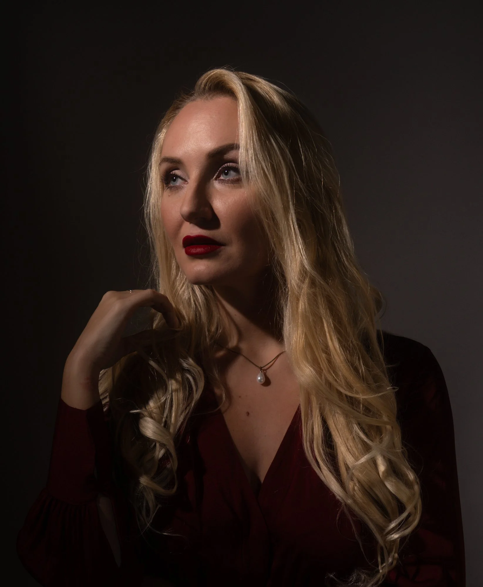 A woman with long blonde hair, red lipstick, and a pearl necklace, looking slightly upward with her hand near her chin against a dark background.