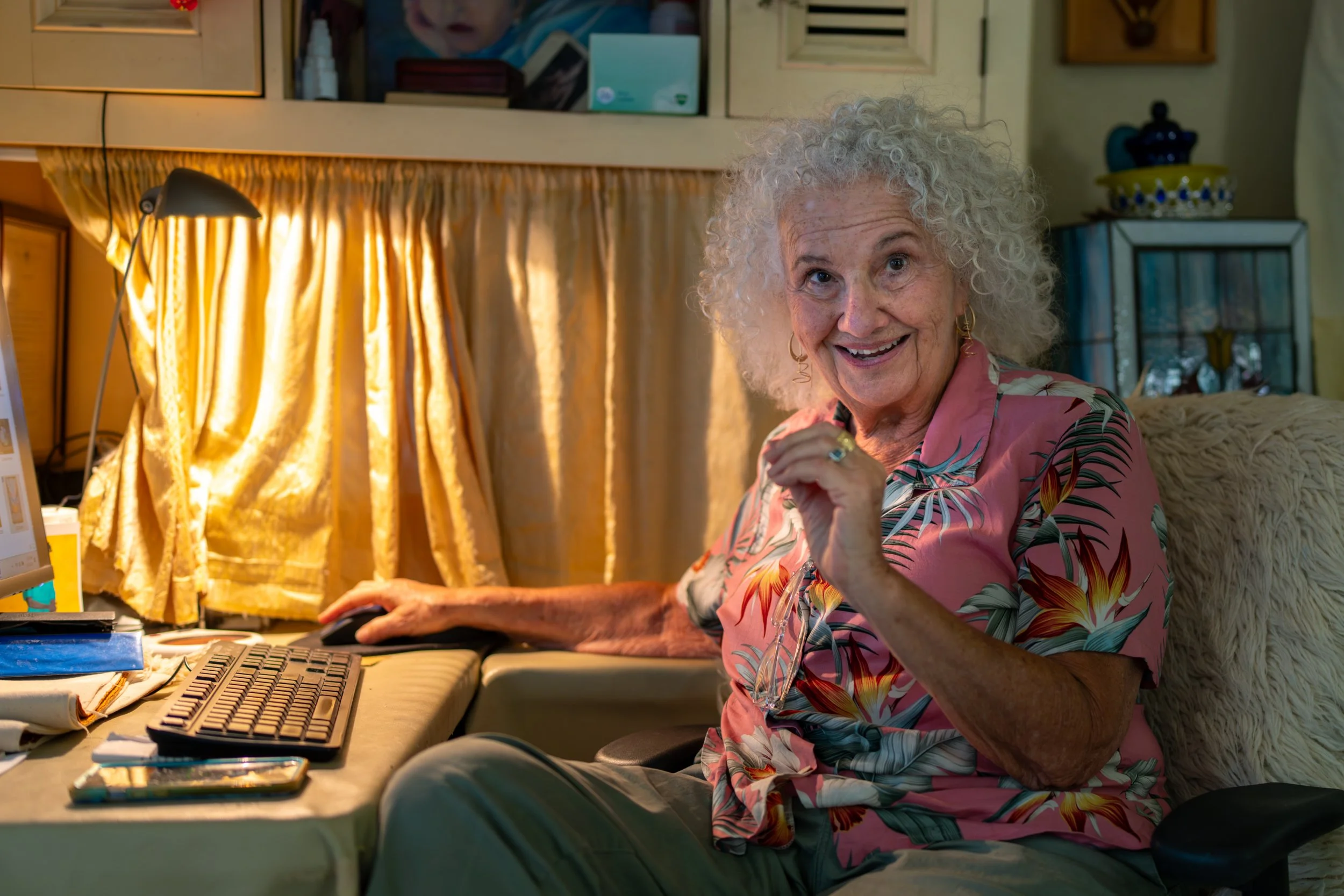 An elderly woman with curly gray hair is sitting at a computer desk, smiling and looking at the camera. She is wearing a pink floral shirt and gold hoop earrings. The desk has a keyboard, mouse, and a smartphone. There is a yellow curtain behind her and various decorative items in the background.