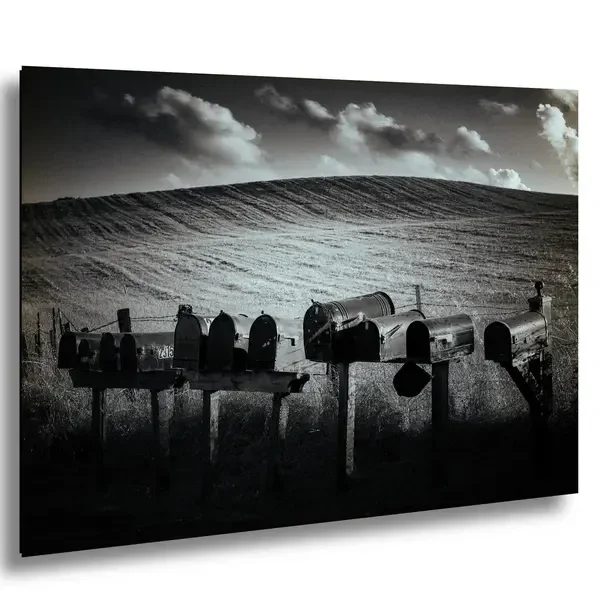 Multiple mailboxes on a rural street with a hilly landscape and cloudy sky in black and white.