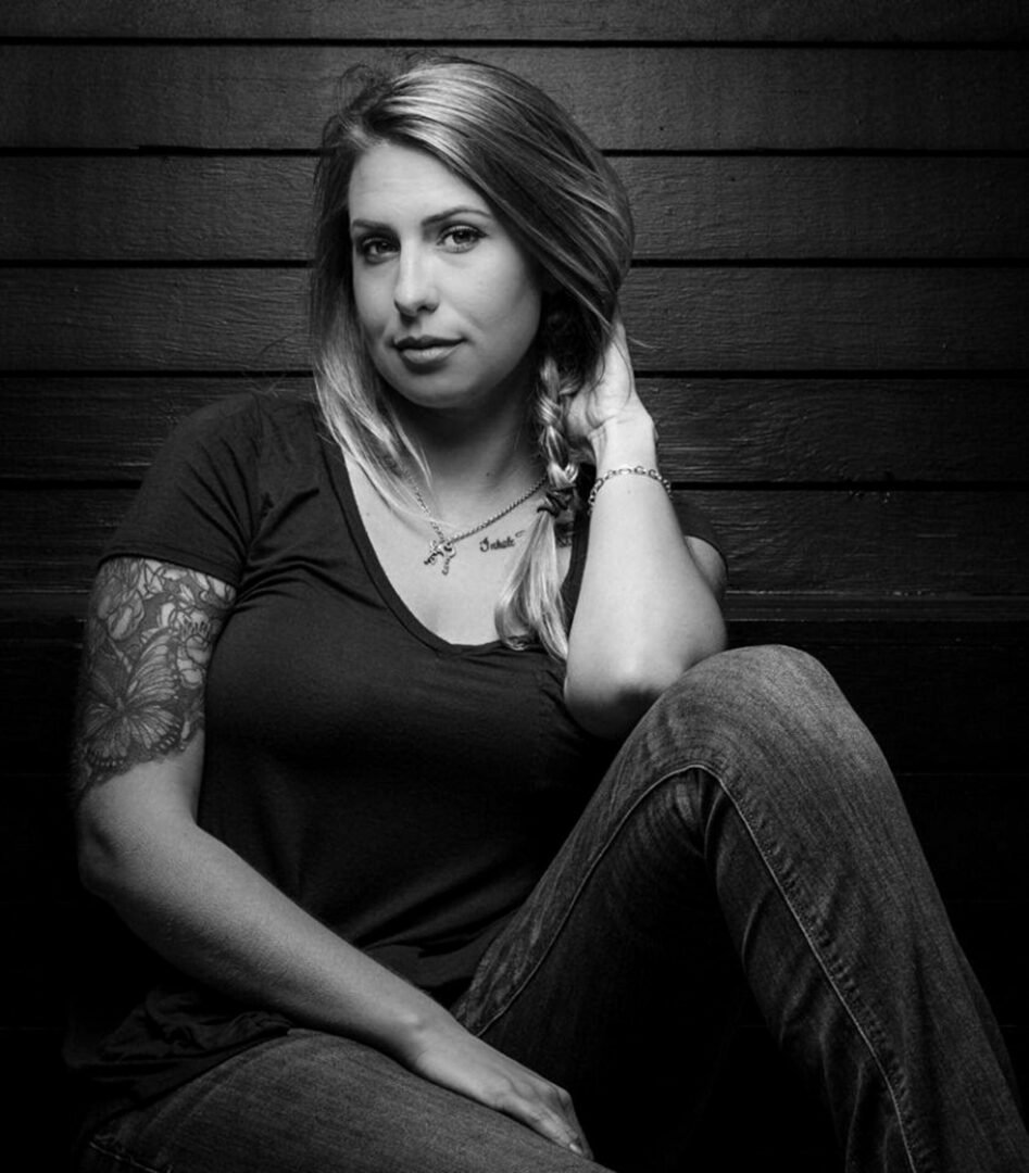 A woman with blonde hair sitting against a wooden wall, wearing a black t-shirt and jeans, with a tattoo on her left arm, and a necklace that says 'Joker'.