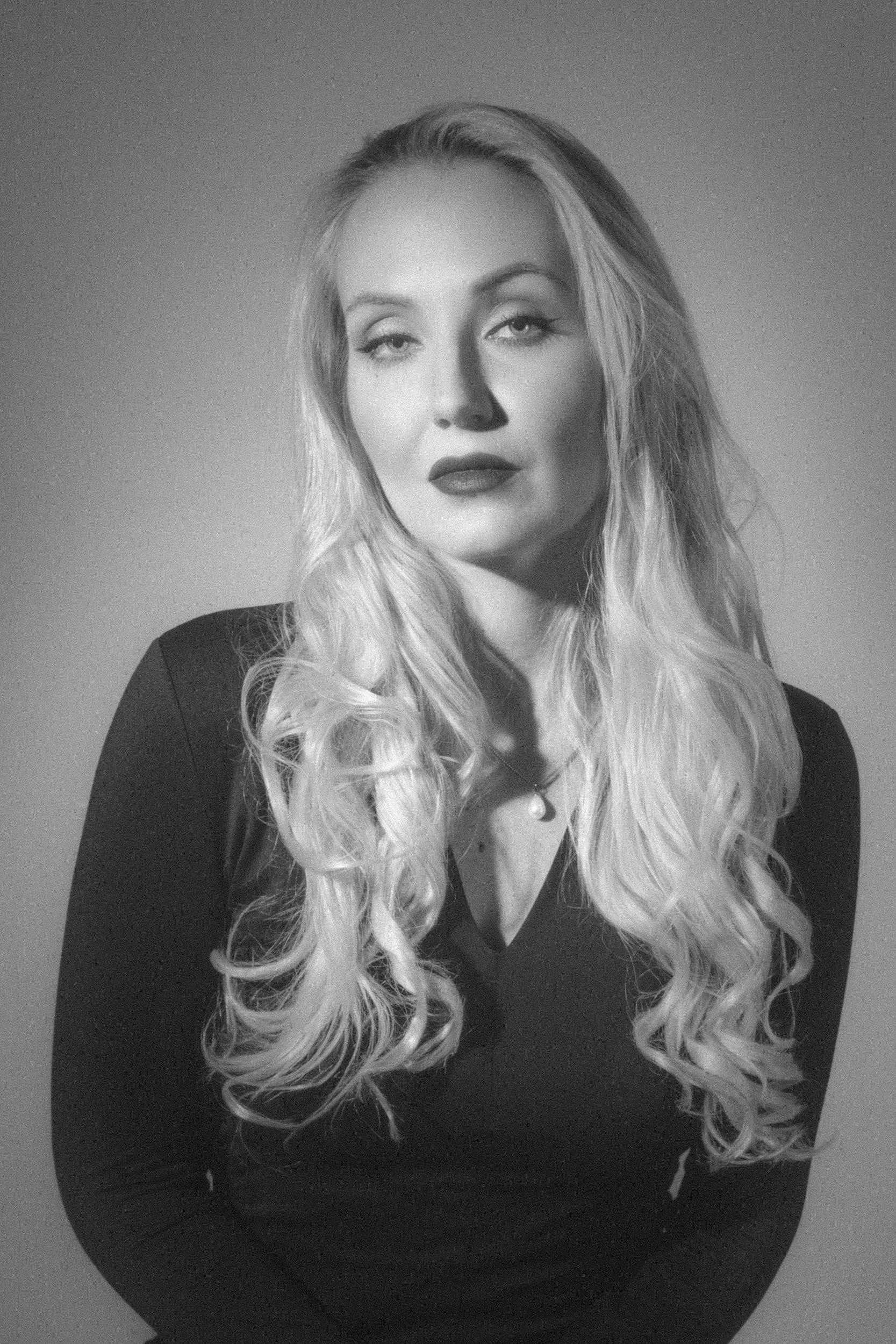 Black and white portrait of a woman with long, wavy blonde hair, wearing a dark top and a necklace with a pearl pendant, against a plain background.