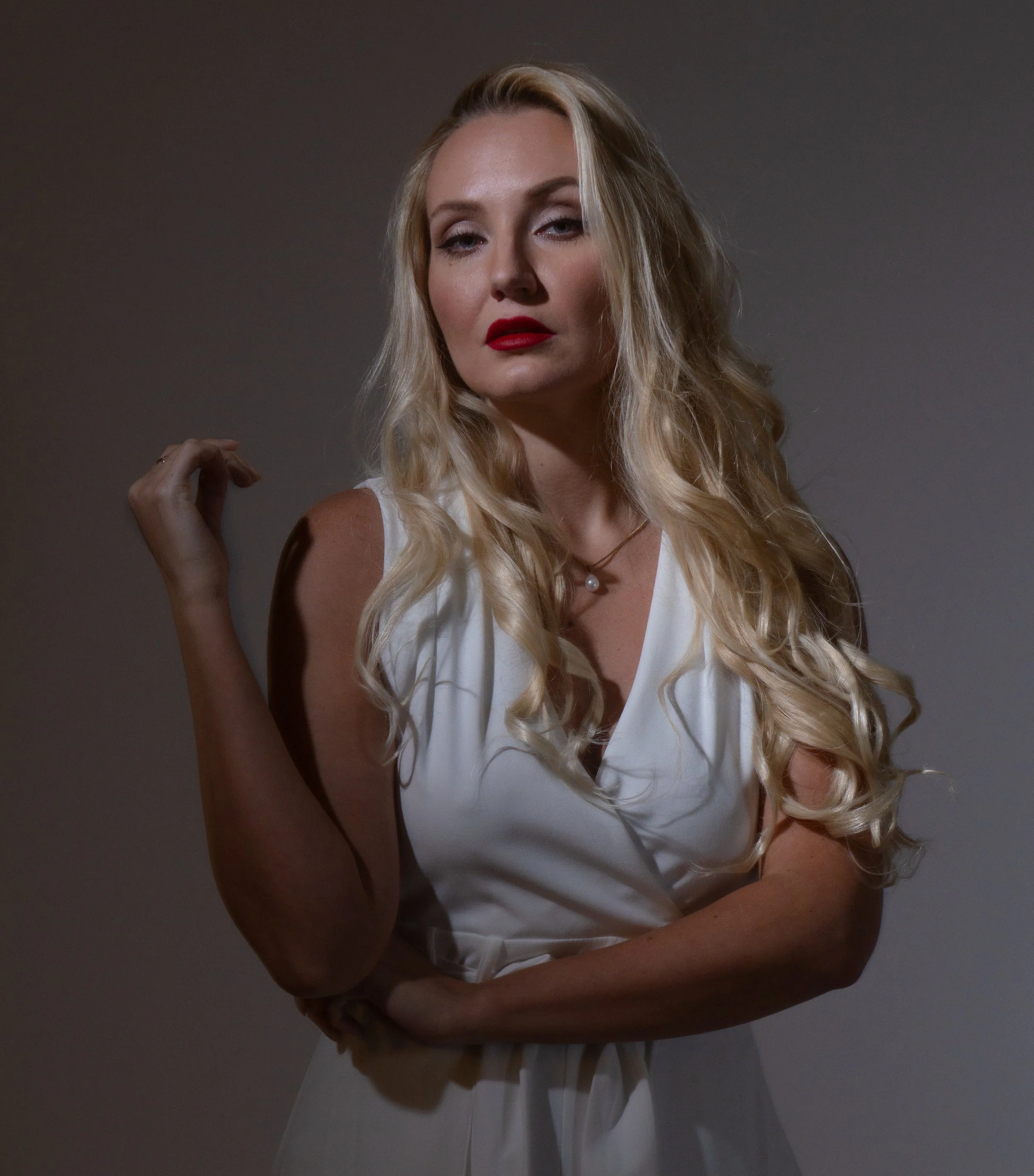 A woman with long blonde curly hair, wearing a white sleeveless top and red lipstick, standing against a plain dark background.