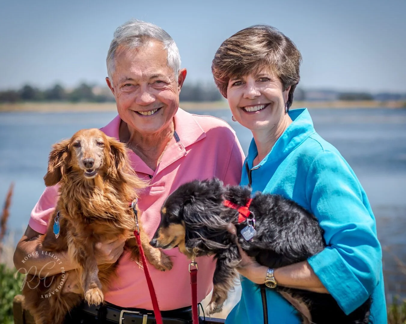 Smiling elderly man and woman standing near water, each holding a dachshund dog outdoors on a sunny day.