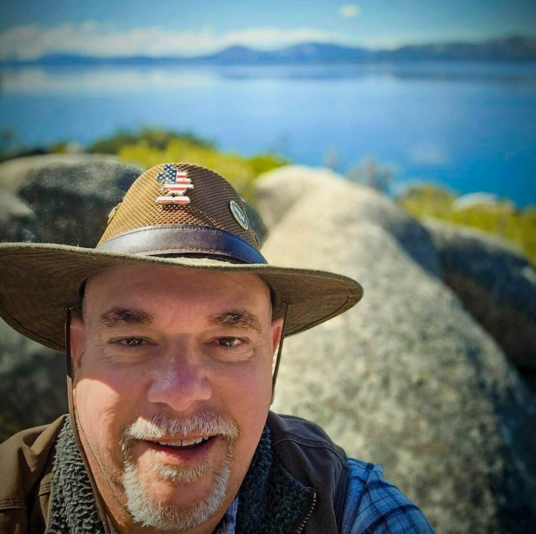 Man smiling outdoors wearing a wide-brimmed hat with an American flag patch, rocks, and a body of water in the background.