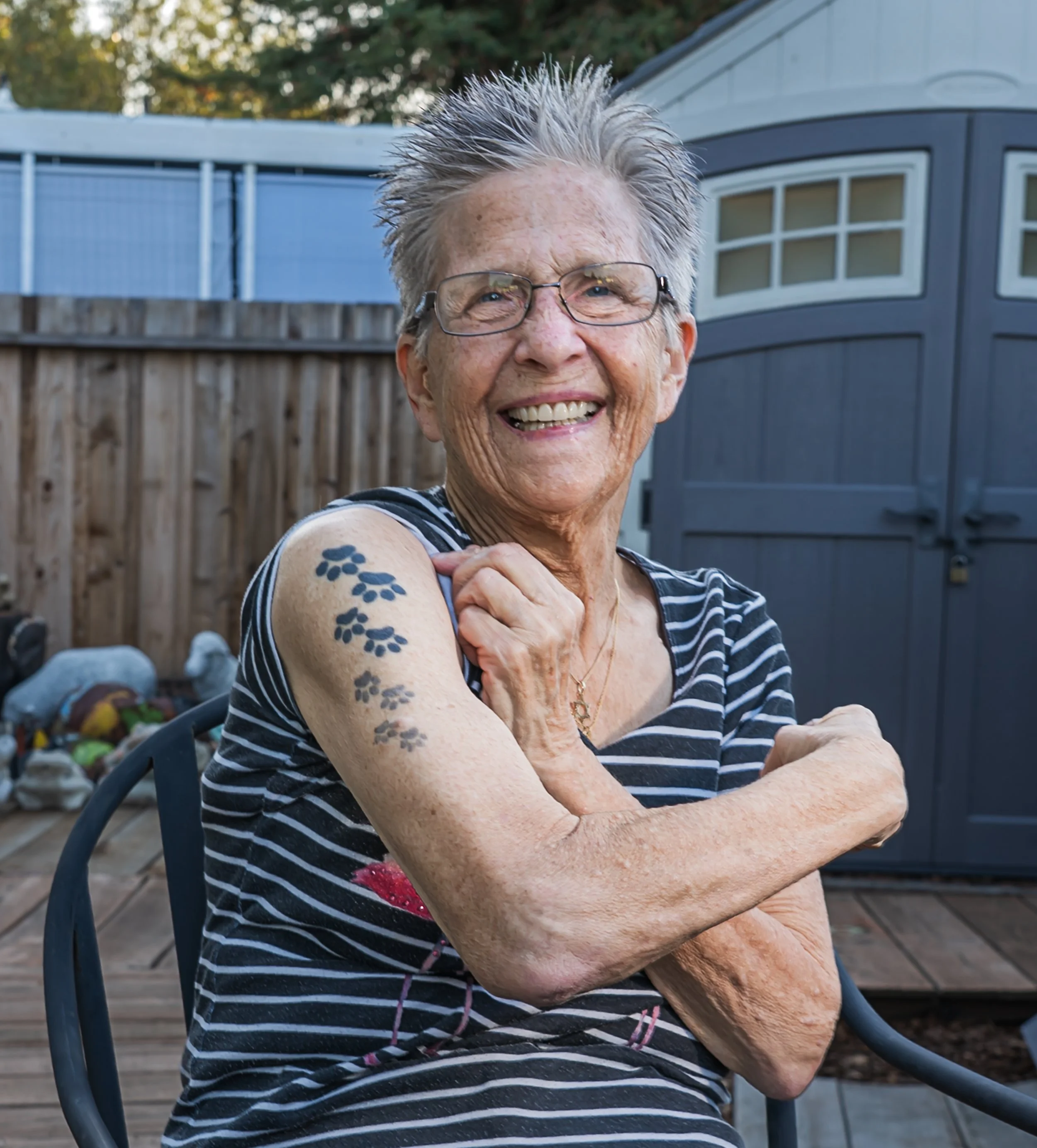 An elderly woman sitting outdoors in a backyard, smiling and flexing her arm to showcase tattoos on her upper arm, wearing glasses and a striped shirt.