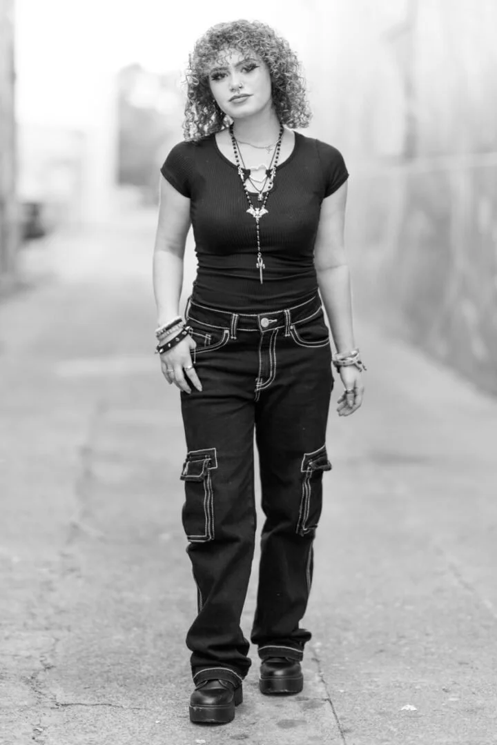 A woman with curly hair walking outdoors in an urban area. She is wearing a black t-shirt, cargo jeans, and platform shoes, accessorized with layered necklaces and bracelets.