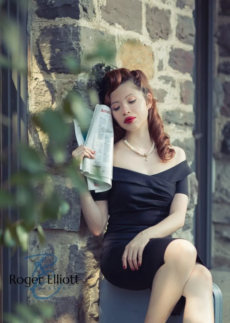 A woman with red hair and red lipstick sits against a stone wall, reading a newspaper. She wears a black off-shoulder dress and a pearl necklace.
