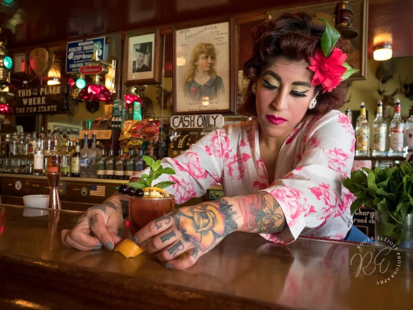 A tattooed woman with vintage style makeup and a pink flower in her hair prepares a cocktail at a bar, with various bottles and framed pictures in the background.