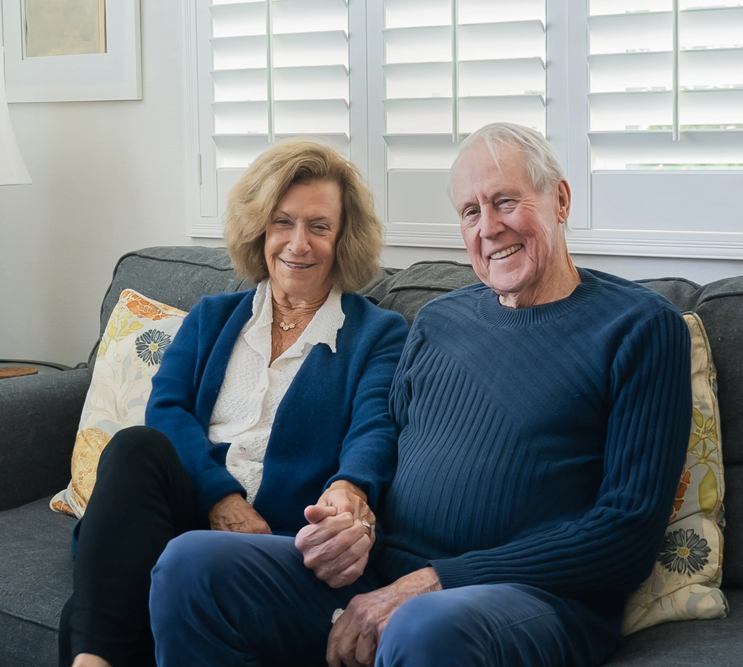 An older couple smiling and holding hands while sitting on a gray couch in a well-lit living room with white shutters behind them.