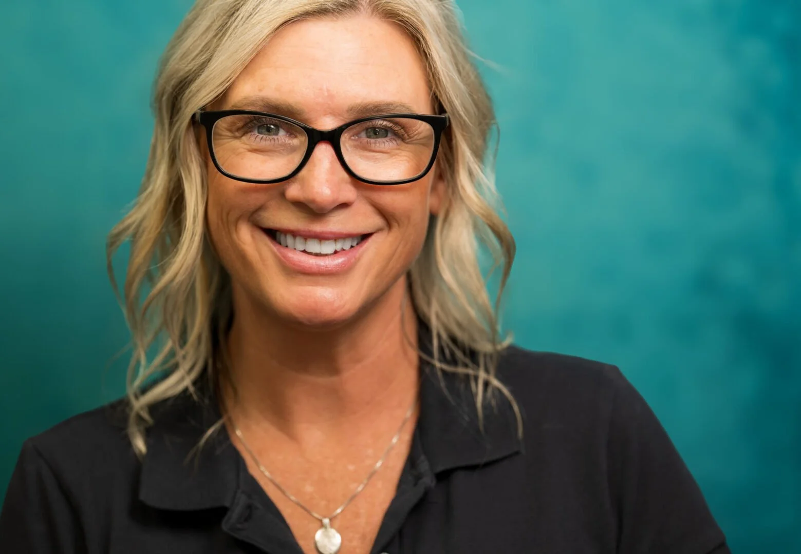A woman with blonde hair wearing glasses, smiling, dressed in a black top, with a turquoise background.