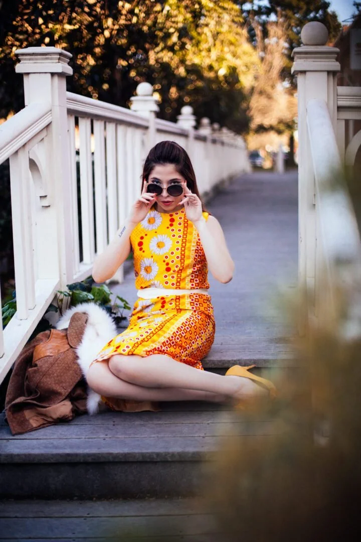 A woman sitting on a wooden sidewalk next to a white picket fence, wearing a yellow dress with floral and geometric patterns, holding sunglasses, with a bag and a dog resting beside her, outdoors during daytime.