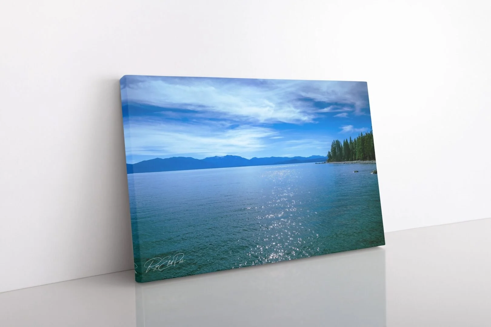 A canvas print of a serene lake with mountains in the distance under a partly cloudy sky, leaning against a white wall.