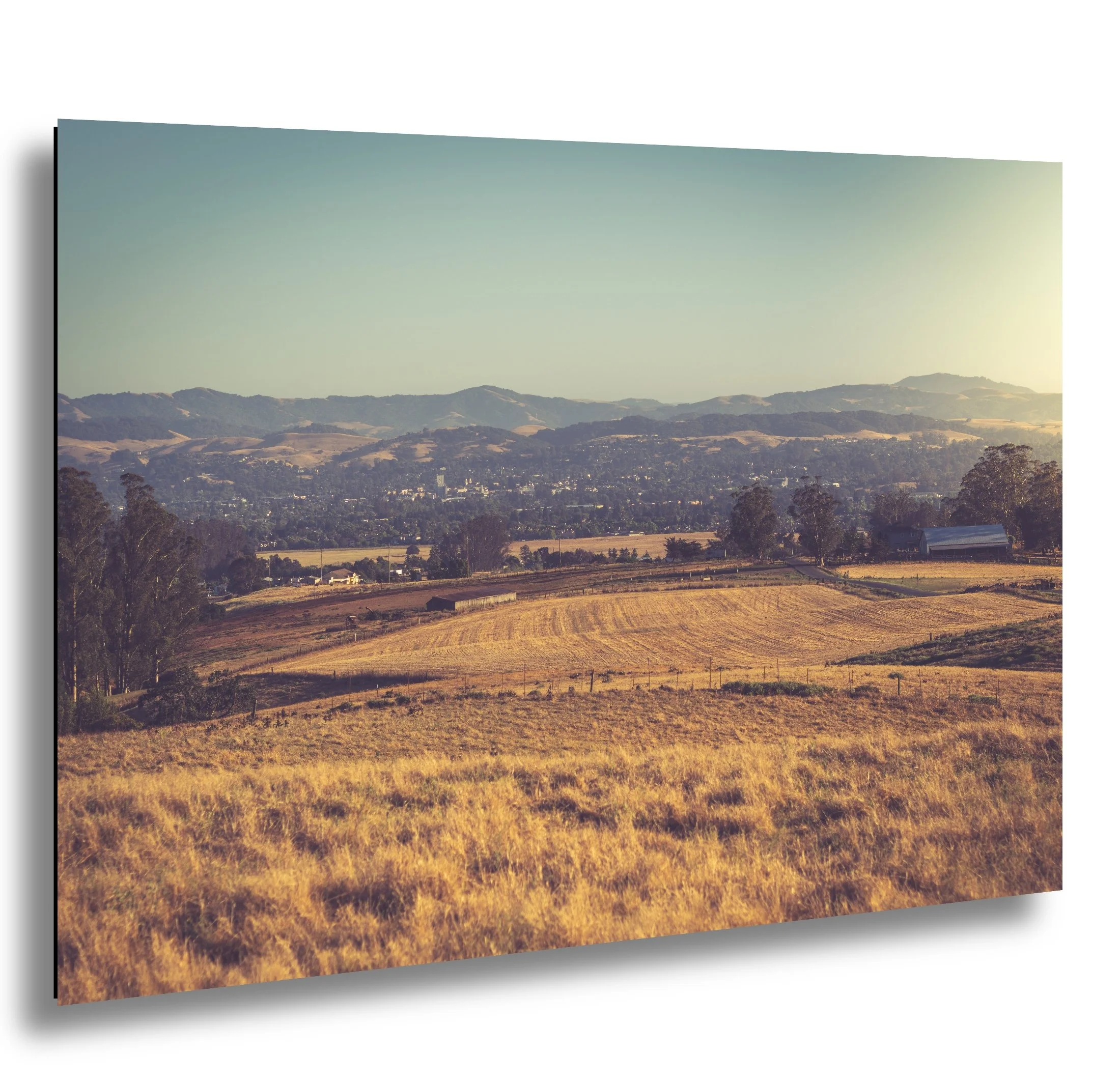 A rural landscape with rolling golden fields, trees, a barn, and distant mountain ranges under a clear sky.