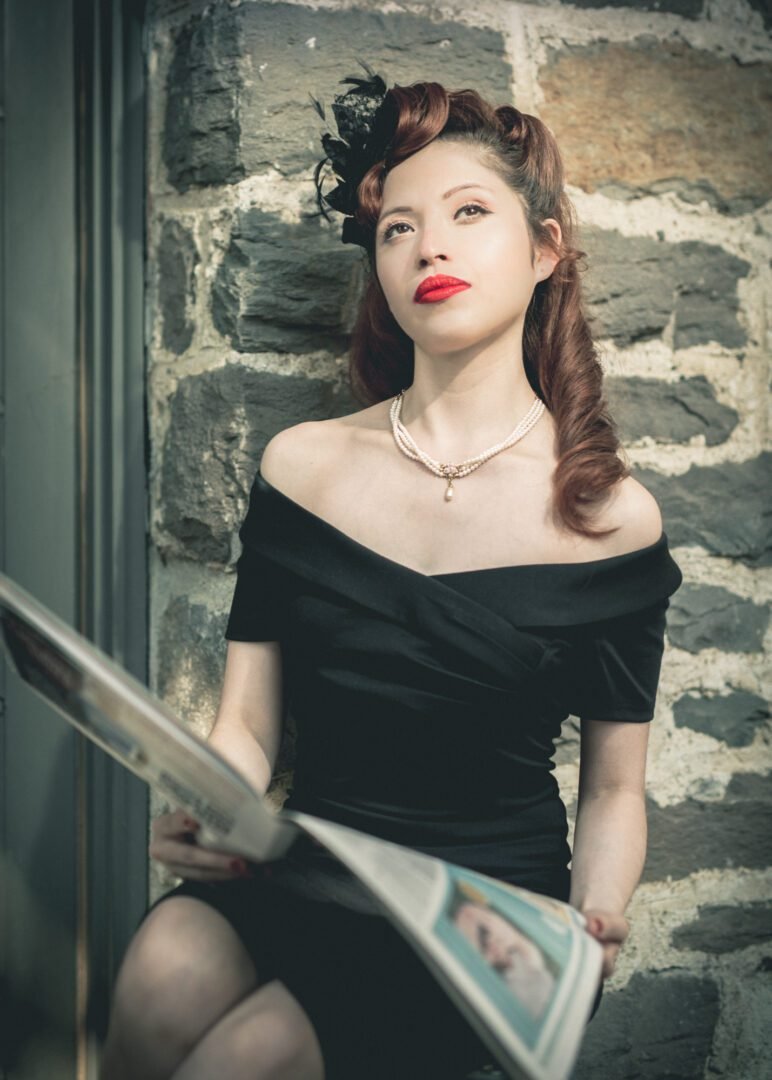 A woman with vintage red hair styled in waves, wearing a black off-shoulder dress, a pearl necklace, and red lipstick, sitting against a stone wall while reading a newspaper.
