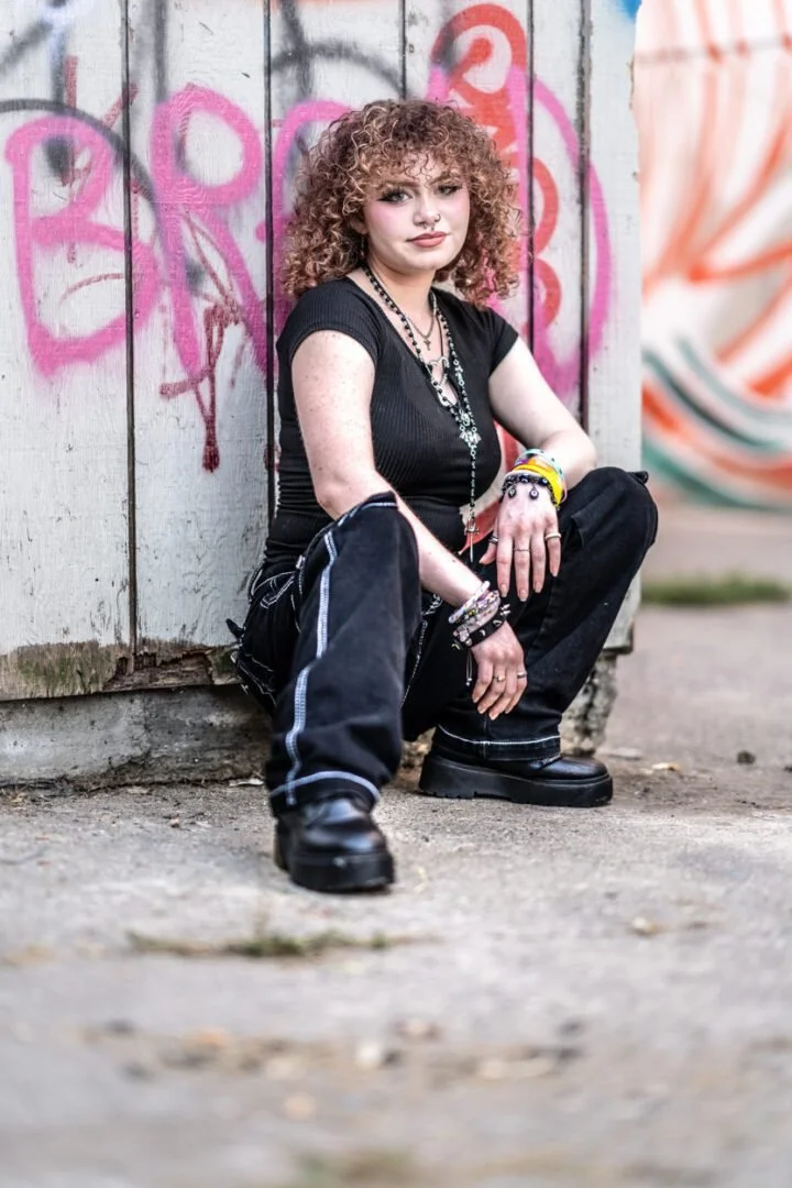 A young woman with curly hair and septum piercing, sitting against a graffiti-covered wooden wall, dressed in black streetwear and accessorized with jewelry.
