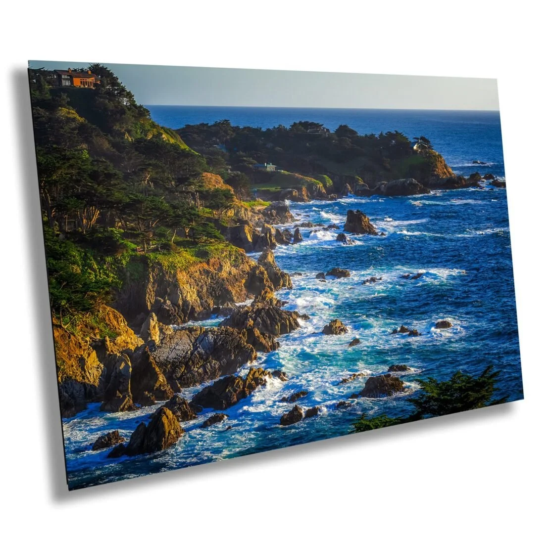 Scenic coastal view with rocky cliffs, green trees, and waves crashing into rocks, overlooking the ocean under clear blue sky.
