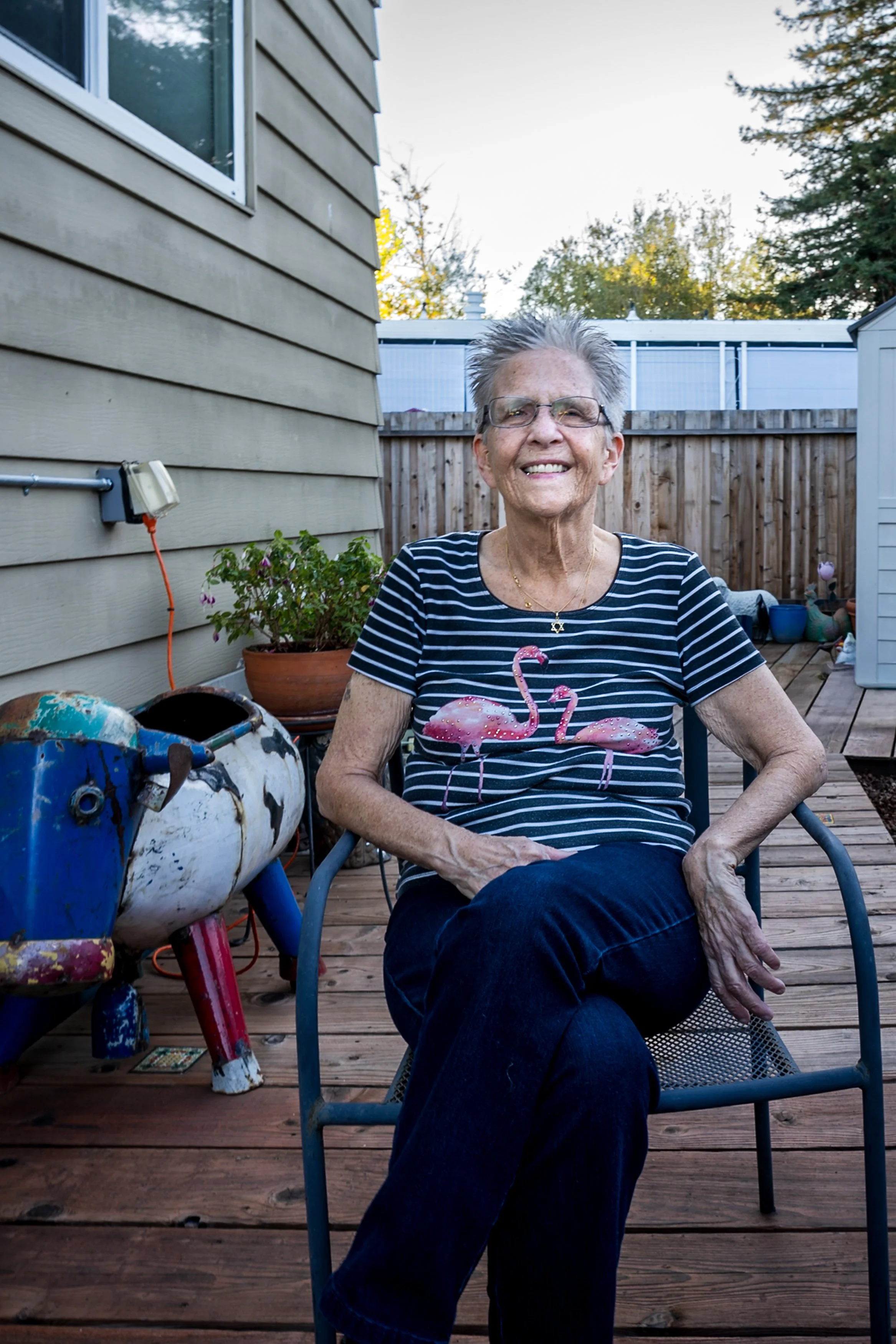An elderly woman with short gray hair and glasses, smiling while sitting on a patio chair. She is wearing a black and white striped shirt with pink flamingos and dark blue jeans. There are plants, a partly visible toy, and a wooden fence in the background.