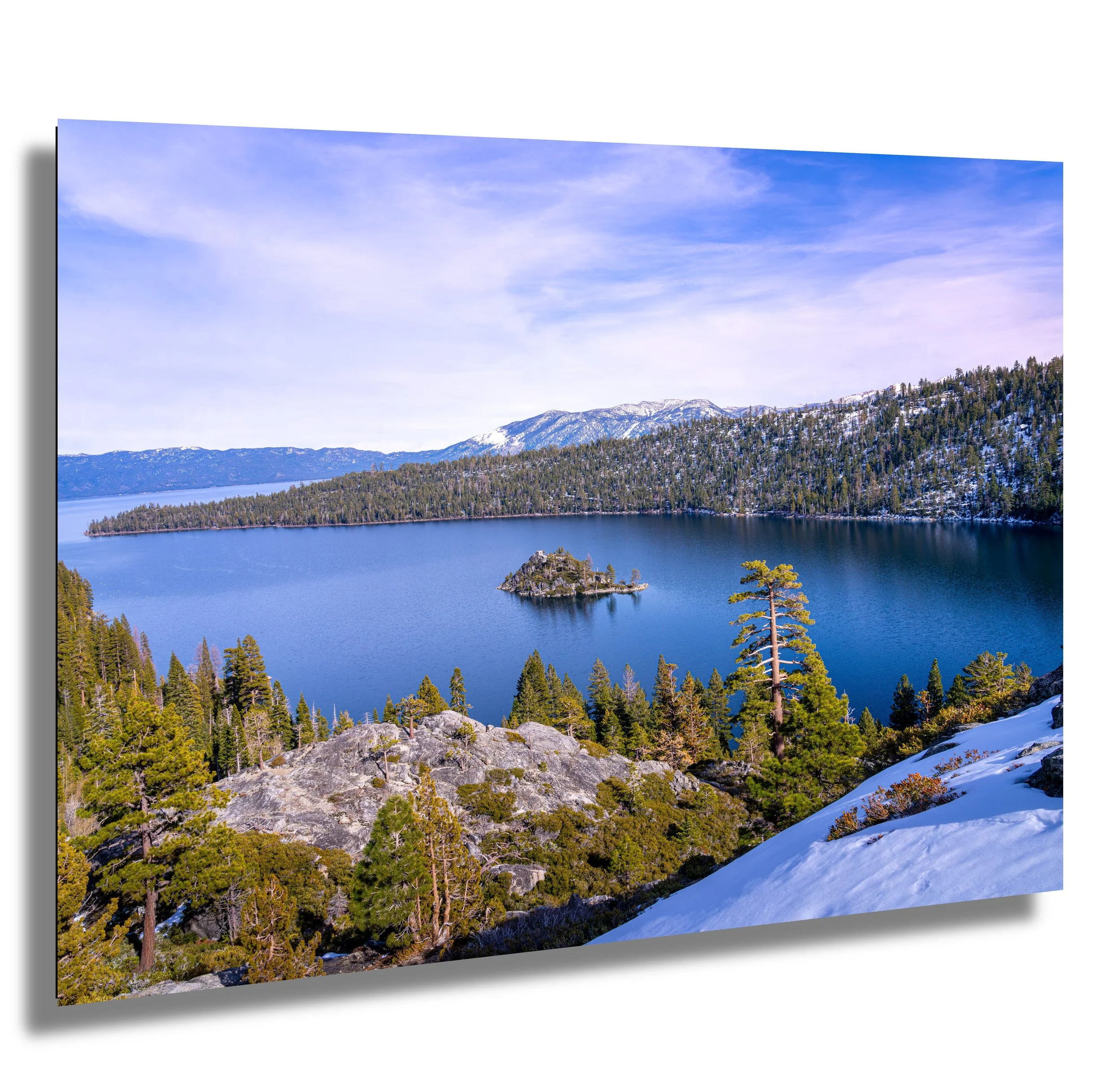 Scenic view of a lake with an island, surrounded by forested mountains with snow, under a partly cloudy sky.