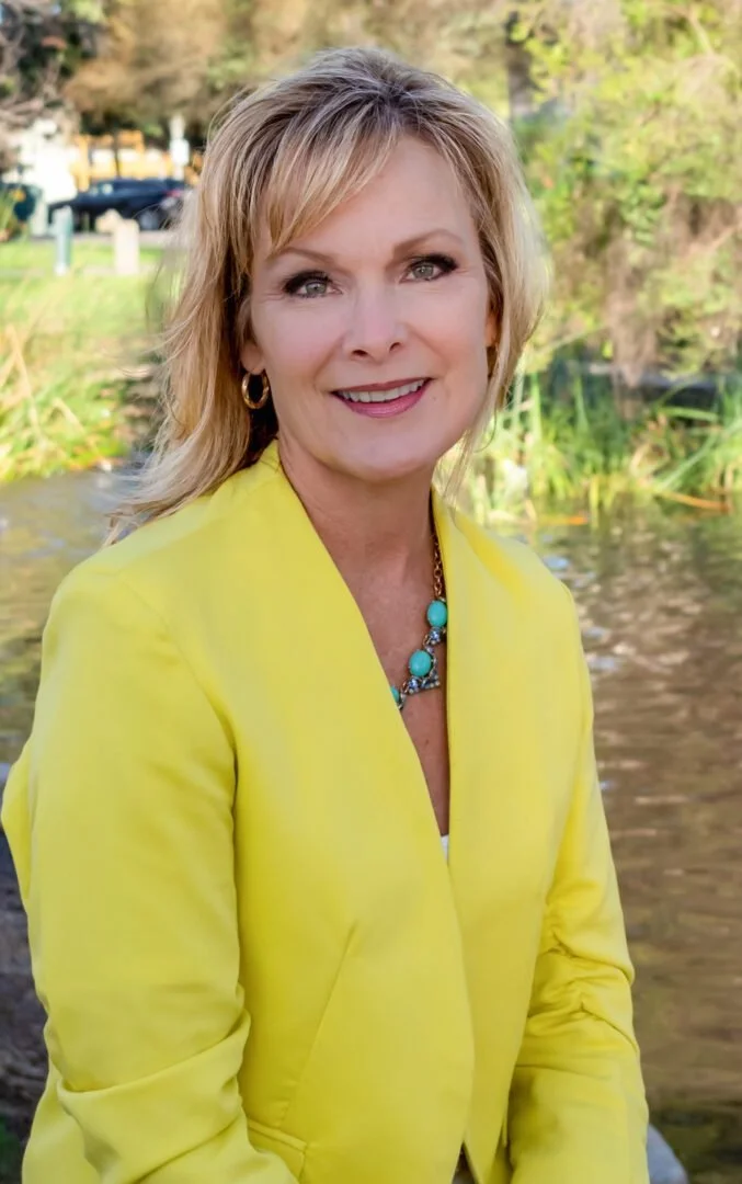 A woman with blonde hair, wearing a bright yellow blazer and turquoise jewelry, standing outdoors near a water body with trees and cars in the background.