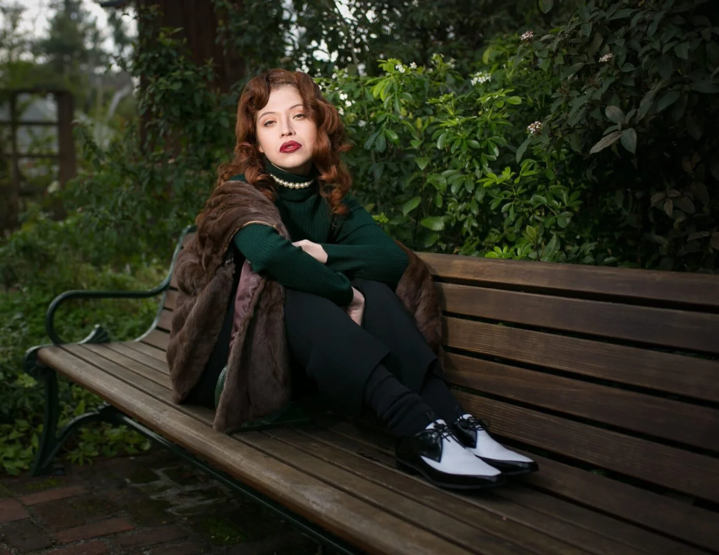 A woman with red hair sitting on a wooden park bench surrounded by green foliage, wearing a dark green top, a pearl necklace, a fur stole, black pants, and black and white shoes.
