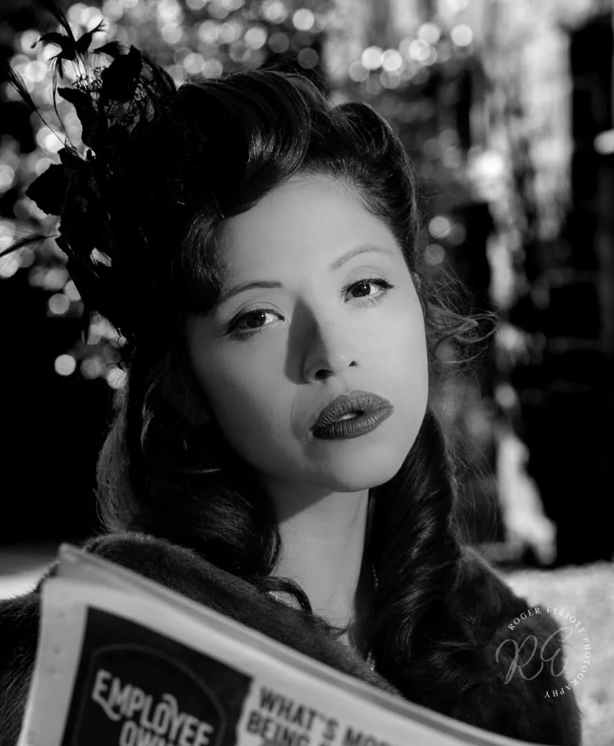 A woman with dark wavy hair styled in vintage fashion, wearing a feathered headpiece, looking directly at the camera, holding a newspaper outdoors.