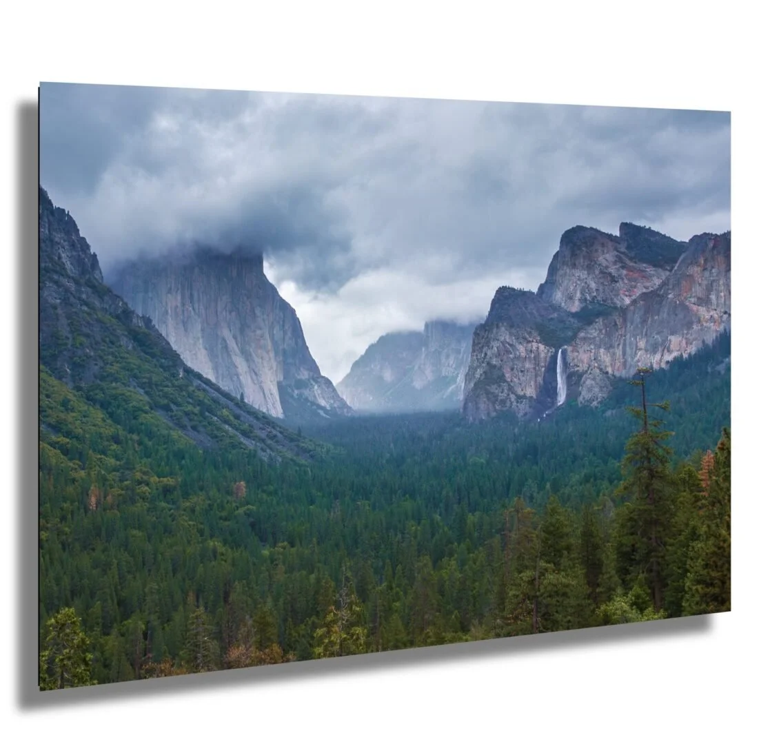Mountain valley with dense forest, cliffs, and waterfall, under cloudy sky.