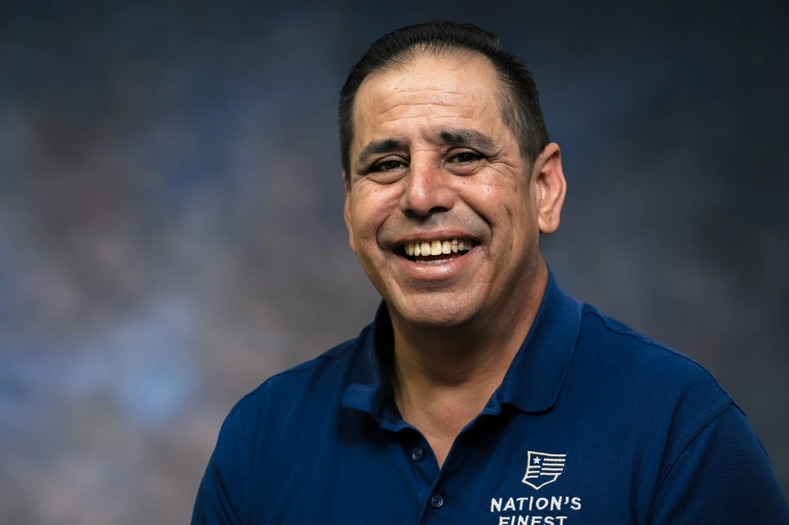 A smiling man with dark hair, wearing a navy blue polo shirt with an American flag logo and the text 'Nation's Finest'.