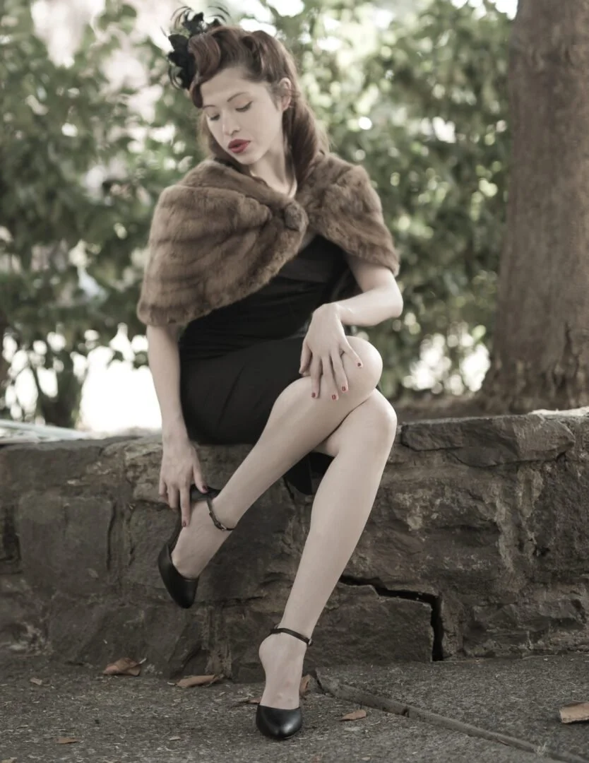 A woman with vintage hairstyle and makeup is sitting on a stone ledge outdoors, wearing a black dress, fur cape, and high heels, with one leg crossed over the other, adjusting her shoe.