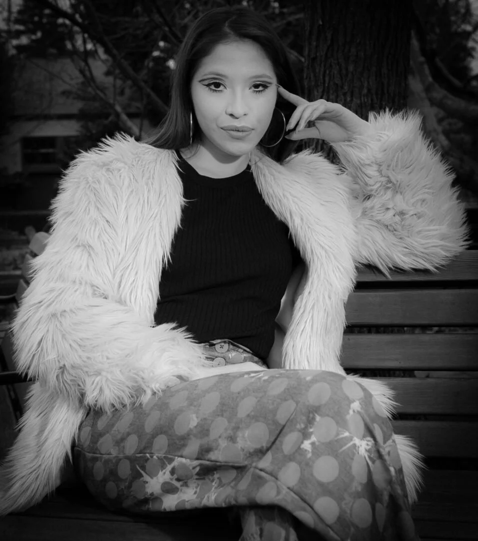 A woman sitting on a wooden bench, wearing a furry coat over a black top, patterned pants, and hoop earrings. She has long hair and is posing with her hand near her face, outdoors with trees in the background.
