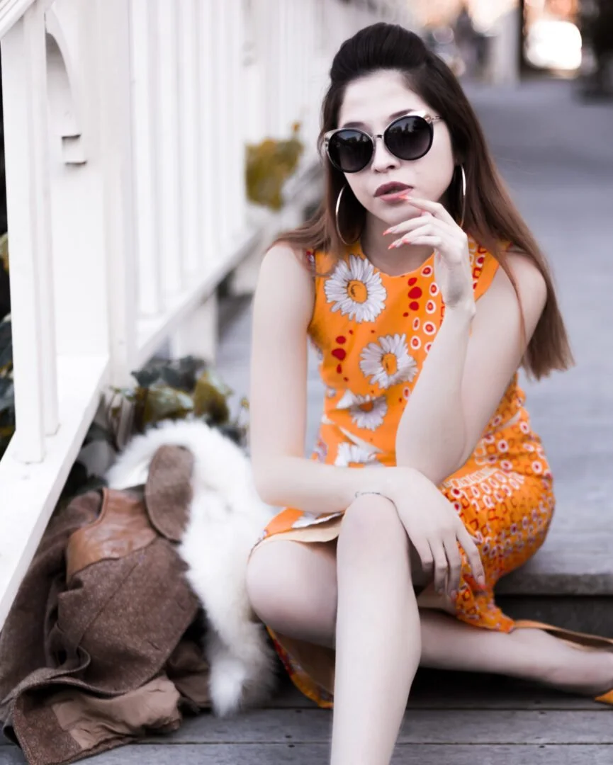 A woman with long hair wearing black sunglasses, hoop earrings, and an orange floral dress sits on a wooden sidewalk next to a white fence. She is touching her lips with her finger and has a brown purse and a white fluffy dog lying beside her.