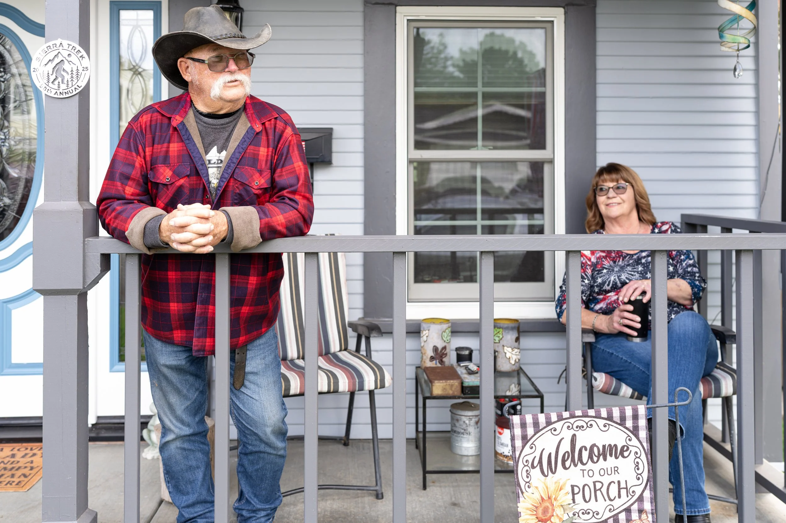 A man and woman sitting on a porch; the man is wearing a cowboy hat and a red plaid shirt, leaning on the railing, while the woman is sitting on a chair holding a drink, wearing glasses and a festive sweater. There is a 'Welcome to our Porch' sign hanging on the porch railing.