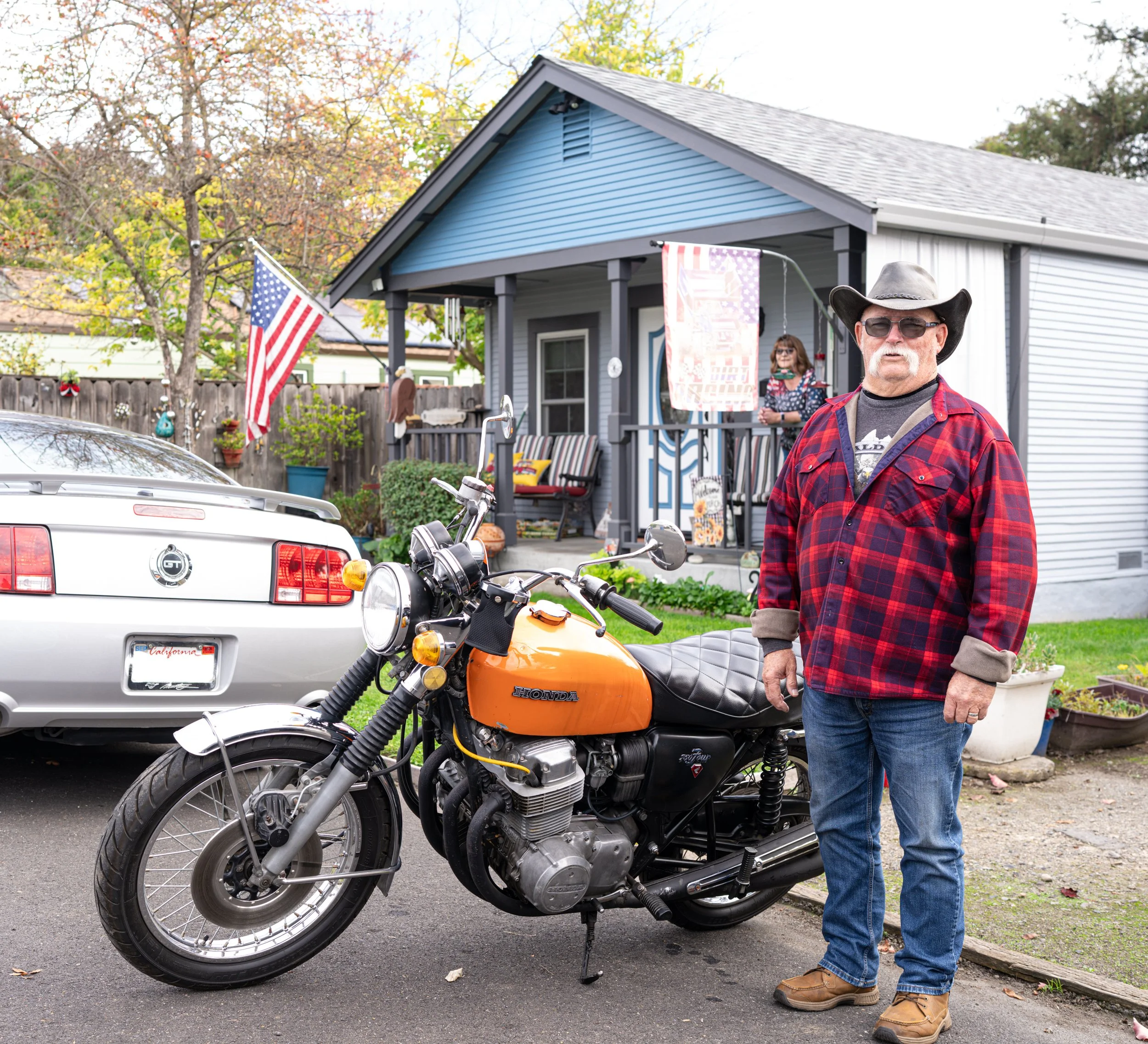 A man in a red plaid shirt and cowboy hat standing next to an orange Honda motorcycle in a residential driveway, with a white car and a blue house in the background.